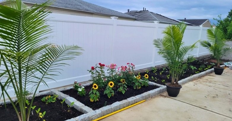 A white fence stands behind vibrant plants and flowers, enhancing the beauty of the landscaped area.