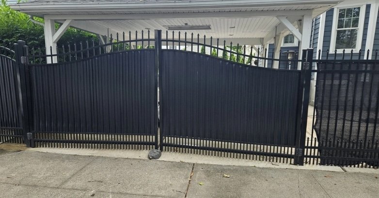 A black wrought iron gate in front of a house, showcasing a sloped driveway leading to the entrance.